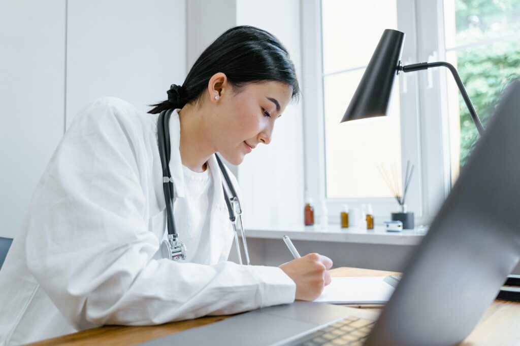 A female doctor writes notes at her desk with a stethoscope around her neck.